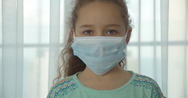 Uma criança em casa em uma máscara médica protetora com um rosto triste e olhos olha para a câmera. Pandemia, covid - 19. — Vídeo de Stock