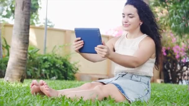 Kobieta używa tabletu ze smartfonem, pisze SMS, komunikuje się z aplikacją, odpoczywa na ulicy, siedzi na trawie w parku. — Wideo stockowe