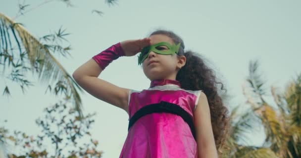 Симпатичная девочка в костюме супергероя, одетая в розовый плащ и зеленую маску героя, машущая вдаль, ребенок играет супергероя на улице, ребенок демонстрирует силу. Супергерой и власть — стоковое видео