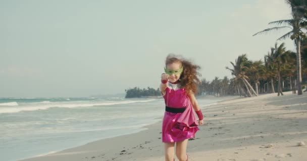 Plays on the Background of the Sea and the Blue Sky and Clouds, Corrects the Green Mask on His Face. Linda niña juega superhéroe. Divertido niño en un impermeable rojo y máscara jugando superhéroe poder. — Vídeo de stock