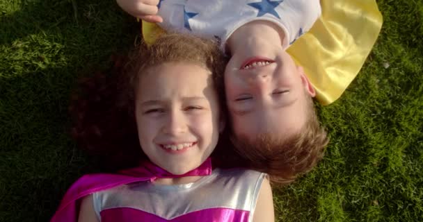 Portrait enfants gais dans un imperméable se reposent, les héros regardent la caméra, sourire, se réjouir. Les enfants super-héros, petite fille et un bébé garçon reposent sur l'herbe en costumes de super-héros. — Video