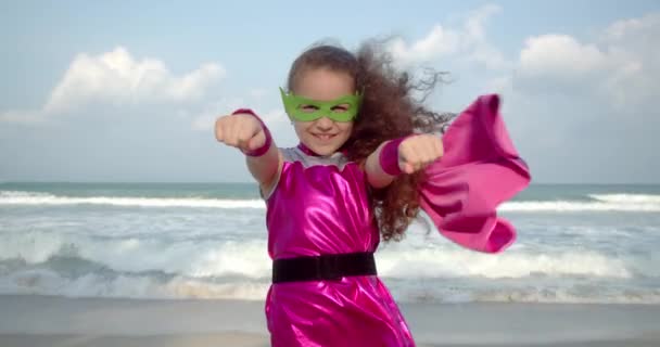 Bebê menina desempenha um super-herói faz movimentos de voo, voa para a frente com a mão levantada para a frente.Criança bonito joga super-herói. Engraçado garoto em uma capa de chuva vermelha e máscara poder de jogo super herói. — Vídeo de Stock