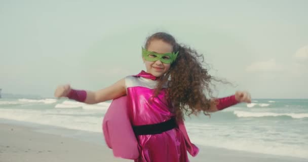 Superhero and power concept.child demonstrates strength.Kid plays a superhero on the Background of Sea and Blue Sky and Clouds.Baby girl in a superhero costume, dressed in a pink cloak and a hero mask. — стоковое видео