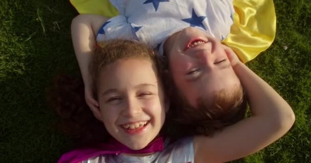 Дети супергерои, маленькая девочка и мальчик лежат на траве в костюмах супергероев. Портрет веселые дети в плаще отдыхают, супергерои смотрят в камеру, улыбаются, радуются. — стоковое видео