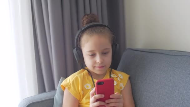Retrato de close-up de uma pequena menina de cabelos encaracolados em fones de ouvido vermelhos. A criança ouve música e canta ao longo da batida. Foco seletivo. Espaço para texto. — Vídeo de Stock