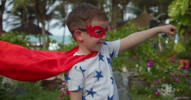 Милый мальчик в костюме супергероя, одетый в красный плащ и маску героя, машущий вдаль, ребенок играет супергероя на улице, ребенок демонстрирует силу. Супергерой и власть — стоковое видео