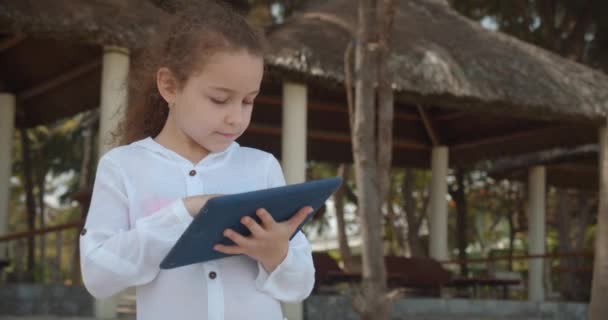 Barnet har en vilostund i tabletten utomhus. Nyfiken söt flicka 7-8 år gammal har kul med en digital surfplatta enhet titta på en tecknad när du går på stranden. — Stockvideo