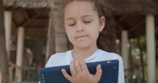 Любознательная симпатичная девочка 7-8 лет развлекается с цифровым планшетным устройством, глядя мультик во время прогулки по пляжу. Ребенок отдыхает играет на планшете на открытом воздухе. — стоковое видео