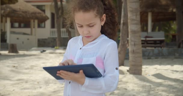 7-8 yaşındaki meraklı kız plajda yürürken dijital tablet cihazıyla eğleniyor. Çocuğun dışarıdaki tablette dinlenme oyunu var.. — Stok video