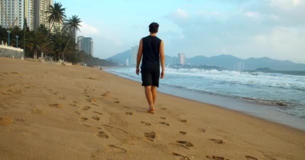 Joven hombre caucásico caminando sobre la arena a lo largo de la orilla al aire libre contra el telón de fondo del hermoso mar.Walk en la arena en la playa al atardecer contra el fondo del mar — Vídeo de stock