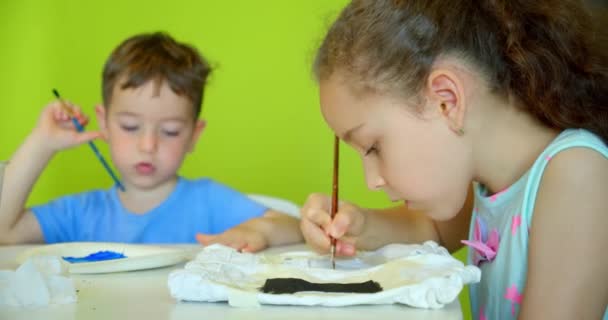 Glada söta barn, en flicka i 7-8 år och förskola pojke, måla hemma med akvareller sitter vid ett vitt bord, liten pojke ritar på papper med färgade färger och baby flicka på hennes vita shorts — Stockvideo
