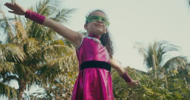 Kid interpreta a un superhéroe al aire libre, lindo niño con un disfraz de superhéroe, vestido con una capa rosa y una máscara de héroe verde, saludando mirando a la distancia. Niño demuestra fuerza. — Vídeo de stock