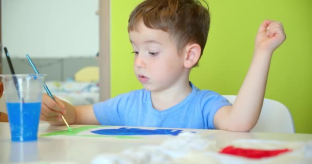 Niños lindos felices, una niña de 7-8 años y un niño preescolar, pintar en casa con acuarelas sentadas en una mesa blanca, niño pequeño dibuja en papel con pinturas de colores y niña en sus pantalones cortos blancos — Vídeos de Stock