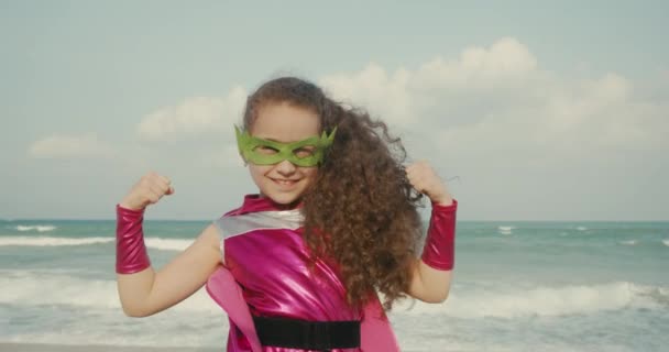 Kid interpreta a un superhéroe en el fondo del mar y el cielo azul y las nubes. Linda niña con un disfraz de superhéroe, vestida con una capa rosa y una máscara de héroe, el niño demuestra fuerza. Superhéroe — Vídeo de stock