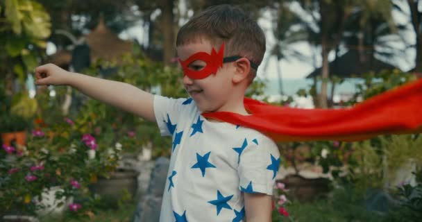 スーパーヒーローの衣装を着たかわいい赤ちゃんの男の子、赤いマントとヒーローマスクを身に着けて、遠くを見て手を振って、子供は屋外でスーパーヒーローを演じ、子供は強さを実証します。スーパーヒーローとパワー — ストック動画