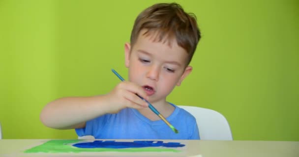 Glückliche süße Kinder, ein Mädchen im Alter von 7-8 Jahren und ein Vorschulkind malen zu Hause mit Aquarellen an einem weißen Tisch sitzend, kleiner Junge zeichnet auf Papier mit bunten Farben und kleines Mädchen auf ihren weißen Shorts — Stockvideo