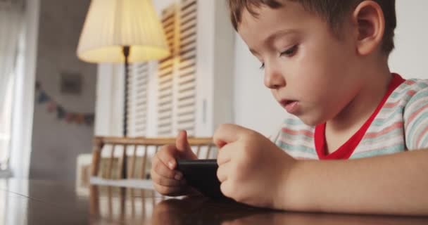 Retrato de um menino, criança com um belo rosto olhando para o telefone enquanto se senta na mesa, uma criança pré-escolar assistindo a um vídeo no telefone joga jogos divertidos. — Vídeo de Stock