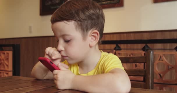 Egy kisfiú portréja, egy gyönyörű arcú gyermek, aki a telefont nézi, miközben az asztalnál ül, egy óvodás gyerek, aki videót néz a telefonon, szórakoztató játékokat játszik.. — Stock videók
