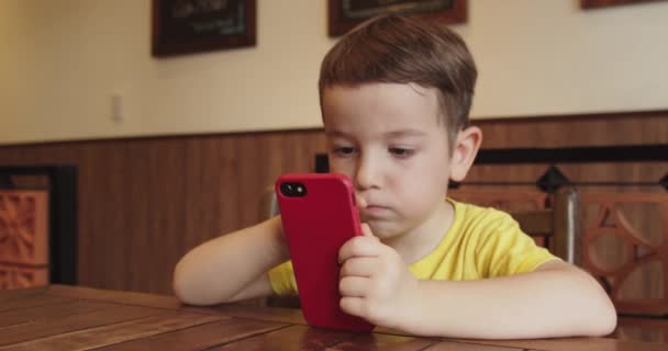 Retrato de un niño pequeño, lindo niño con una mirada en el teléfono mientras está sentado en la mesa, un niño preescolar viendo un video en el teléfono juega juegos entretenidos. — Vídeo de stock