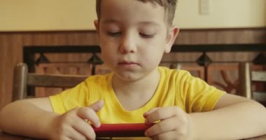 Küçük bir çocuğun portresi, masada otururken telefona bakan sevimli bir çocuk, telefonda video izleyen bir anaokulu çocuğu, eğlenceli oyunlar oynuyor..