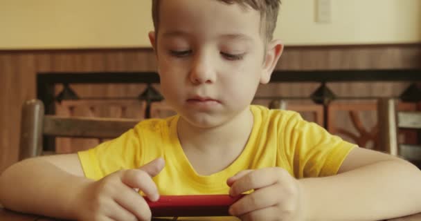 Retrato de un niño pequeño, lindo niño con una mirada en el teléfono mientras está sentado en la mesa, un niño preescolar viendo un video en el teléfono juega juegos entretenidos. — Vídeo de stock