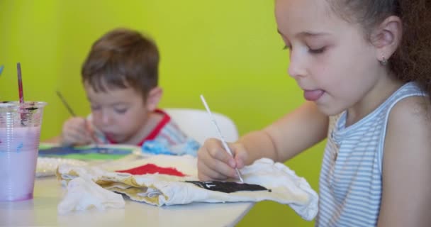 Crianças bonitos felizes, uma menina de 7-8 anos de idade e menino pré-escolar, pintar em casa com aquarelas sentadas em uma mesa branca, menino desenha em papel com tintas coloridas e bebê menina em seus shorts brancos — Vídeo de Stock