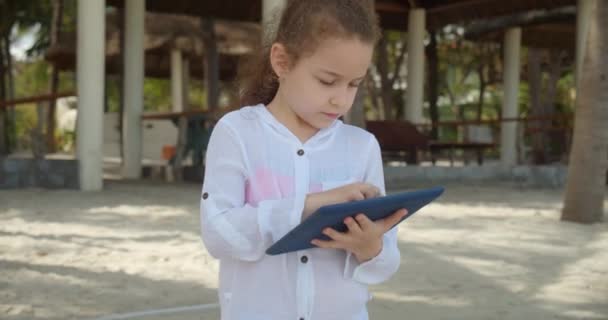 Nyfiken söt flicka 7-8 år gammal har kul med en digital surfplatta enhet titta på en tecknad medan du går på stranden. Barnet har en vilostund i tabletten utomhus. — Stockvideo