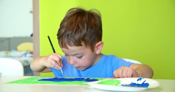 Mutlu şirin çocuklar, 7-8 yaşlarında bir kız çocuğu ve anaokulu çocuğu, beyaz bir masada oturan suluboya boyayla evde resim yapıyor, küçük çocuk renkli boyalar çiziyor ve beyaz şortuna küçük kız çocuğu çiziyor. — Stok video