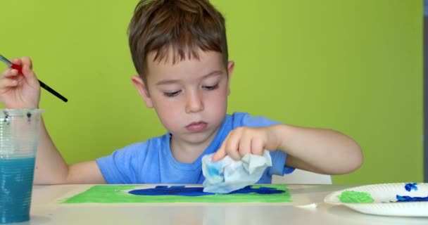 Glückliche süße Kinder, ein Mädchen im Alter von 7-8 Jahren und ein Vorschulkind malen zu Hause mit Aquarellen an einem weißen Tisch sitzend, kleiner Junge zeichnet auf Papier mit bunten Farben und kleines Mädchen auf ihren weißen Shorts — Stockvideo