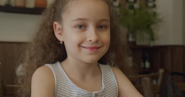 Portrait de petite fille drôle, enfant souriant, regardant la caméra, assis à la table à la maison, enfant mignon avec joli visage. Concept d'enfance heureuse. — Video