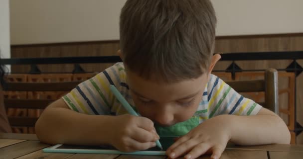 Portrait d'un enfant assis à une table à la maison et dessiné sur sa tablette avec un stylo spécial, un enfant d'âge préscolaire utilise une tablette spéciale pour le dessin. — Video