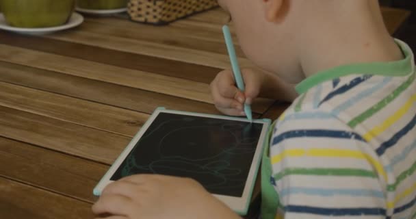 Un retrato de un niño se sienta en una mesa en casa y dibuja en su tableta con una pluma especial, un niño en edad preescolar utiliza una tableta especial para dibujar. — Vídeos de Stock