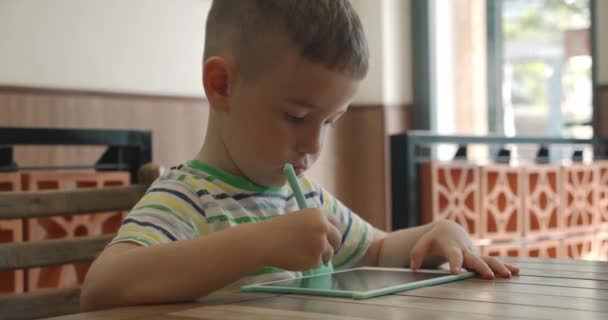 Ein Kind sitzt zu Hause an einem Tisch und zeichnet mit einem speziellen Stift auf seinem Tablet, ein Vorschulkind zeichnet mit einem speziellen Tablet. — Stockvideo