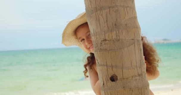 Счастливая маленькая девочка на пляже в купальнике и большой шляпе смотрит из-под пальмы, смотрит в камеру, счастливо улыбается. Ребенок, дети, эмоции. — стоковое видео