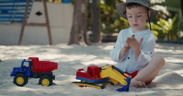 Bébé garçon mignon assis seul sur la plage joue avec une grue en plastique et une voiture sur le sable sur la plage. portraits en gros plan d'enfants et de mains d'enfants jouant avec du sable sur la plage, jouets en plastique — Video