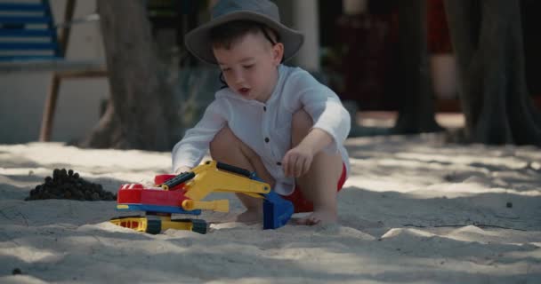 かわいい赤ちゃんの男の子がビーチで砂の中にプラスチック製の車で遊んでいます。ビーチで砂と遊ぶ子供たちの手のクローズアップ、プラスチックのおもちゃ. — ストック動画
