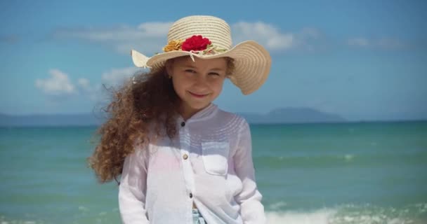 Portræt af en sjov lille pige i en hat, et smilende barn kigger på kameraet, står på stranden ved havet, et sødt barn med et smukt ansigt står på stranden. Happy barndom koncept. – Stock-video