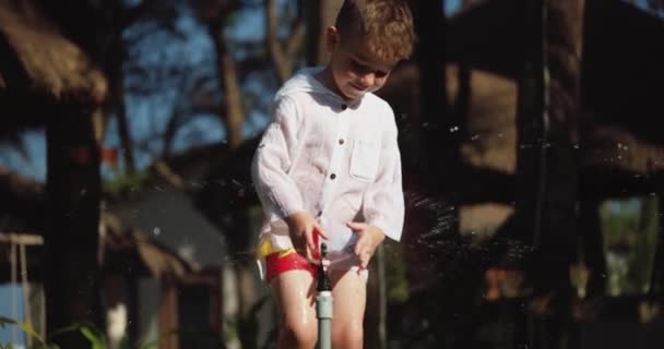 Мила щаслива дитина грає з садовим спринклером в літній спекотний день. Маленький хлопчик із задоволенням стрибає на задньому дворі під розпилювачем води з шланга. . — стокове відео