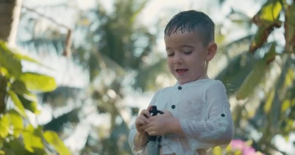 Leuk vrolijk kind spelen met tuin sprinkler op de zomer hete dag. Kid hebben plezier op de achtertuin springen onder spuiten water van slangen.Little jongen gelukkige jeugd. — Stockvideo