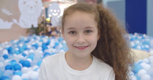 Retrato de uma menina alegre, criança sorridente, salta de bolas de plástico das crianças, criança olha para a câmera, uma linda menina com um belo jogo de rosto em casa, se alegra. Conceito de infância feliz — Vídeo de Stock