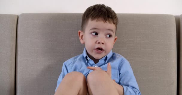 Retrato con una cara misteriosa de un niño pequeño, niño mira a un lado, sentado en el sofá en casa, lindo niño preescolar con una cara hermosa, sonriendo, sentado en el sofá. Concepto de infancia feliz. — Vídeos de Stock