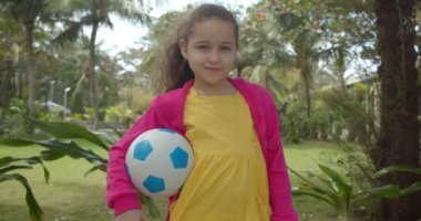 Elinde futbol topu olan sevimli küçük bir kız kameraya bakıp gülümsüyor ve top oynamaya hazırlanıyor palmiye ağaçları, ağaçlar ve güneş ışığına karşı..