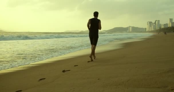 A fiatal kaukázusi férfi szabadtéri futással gyakorol, gyönyörű tengeri háttérrel. Kocogni a homokon a parton naplementekor a gyönyörű tenger hátterében, nagy, gyönyörű hullámok.. — Stock videók