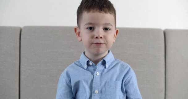 Słodki chłopczyk patrzy w kamerę, siedzi na kanapie w domu i je batonik czekoladowy. Szczęśliwego dzieciństwa. — Wideo stockowe