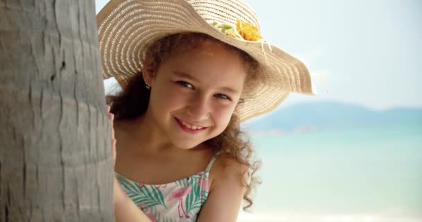 Şapkalı komik küçük bir kızın portresi, kameraya bakan gülümseyen bir çocuk, deniz kıyısında duruyor, güzel yüzlü şirin bir çocuk sahilde duruyor. Mutlu çocukluk kavramı. — Stok video