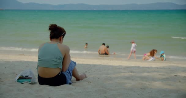 Kobieta siedzi na piasku na plaży, wysyłając SMS-a przez telefon komórkowy, jej rodzina, dzieci i mąż pływają w morzu niedaleko brzegu.. — Wideo stockowe