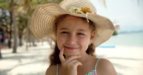 Portrait d'une petite fille drôle dans un chapeau, un enfant souriant regardant la caméra, se tient sur la plage au bord de la mer, un enfant mignon avec un beau visage se tient sur la plage. Concept d'enfance heureuse. — Video