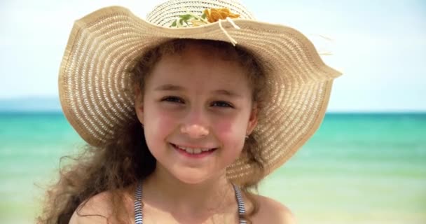 Retrato de una niña divertida en un sombrero, un niño sonriente mirando a la cámara, se encuentra en la playa junto al mar, un niño lindo con una cara hermosa se encuentra en la playa. Concepto de infancia feliz. — Vídeos de Stock
