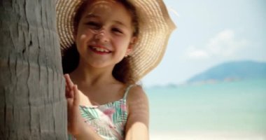 Şapkalı komik küçük bir kızın portresi, kameraya bakan gülümseyen bir çocuk, deniz kıyısında duruyor, güzel yüzlü şirin bir çocuk sahilde duruyor. Mutlu çocukluk kavramı.