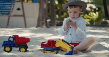 Plastik bir vinç ve kumsalda kumsalda araba ile oynayan bir çocuk. Plajda kumla oynayan çocuğun yakın plan portresi, plastik oyuncaklar..